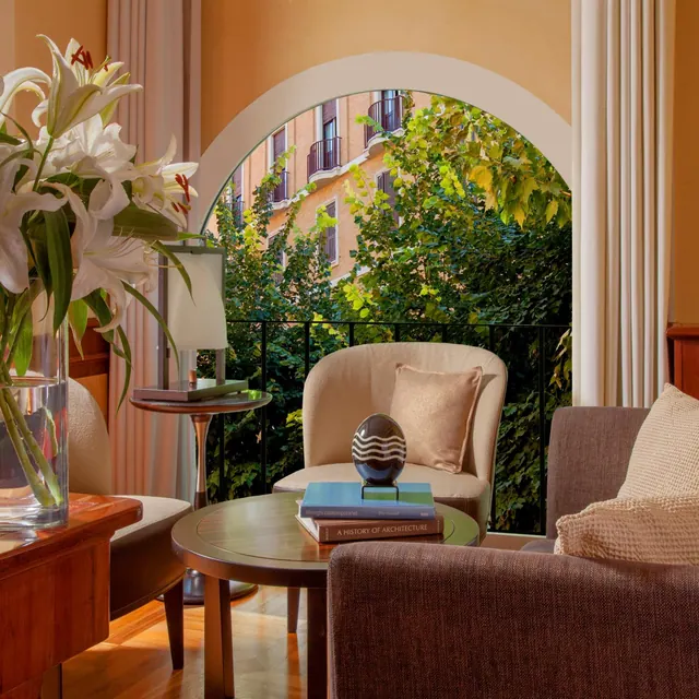 A seating area with armchairs, a round coffee table holding books and a glass sculpture, and an arched window looking out onto green trees and a brick building with balconies.