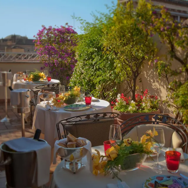 An outdoor rooftop dining terrace with set tables, potted flowering plants, a wine bucket and a bread basket in sunlight, with rooftops visible behind.