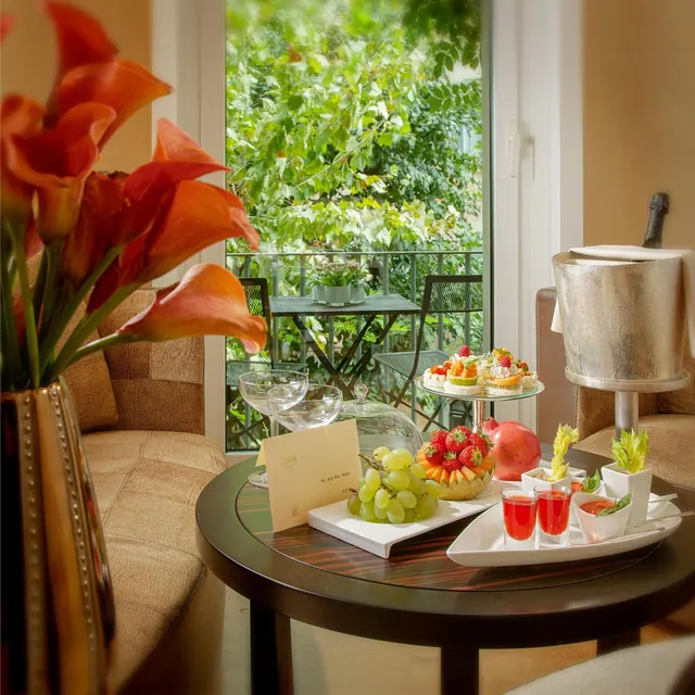 A round dark wooden table holds fruit, cakes, juice shots and a champagne bucket beside a sofa, with a balcony and greenery visible through the open glass door.