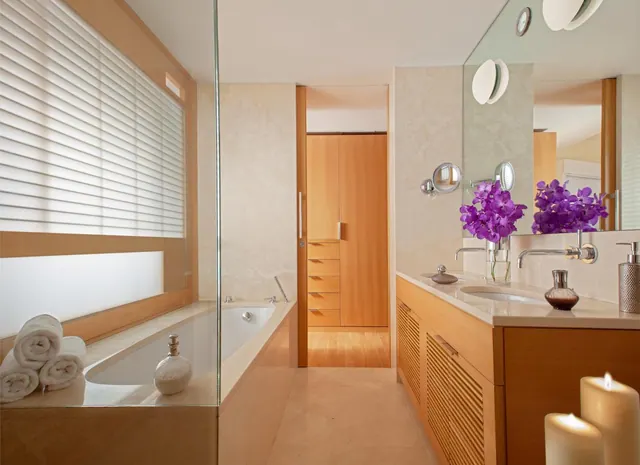 A bathroom with a built-in bathtub beside a blinded window, a wooden double vanity with two basins and purple orchids, and a wooden wardrobe in the background.
