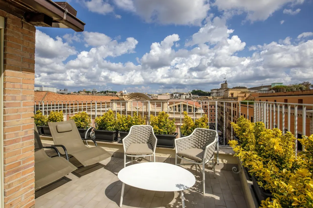 A rooftop terrace with lounge chairs, a round white table and potted shrubs overlooks rooftops and buildings under a cloudy blue sky.