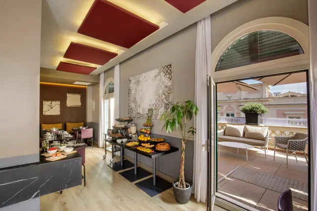 A breakfast room with a fruit and pastry buffet on a dark table, red ceiling panels, and an open arched door onto a terrace with a sofa and chairs.