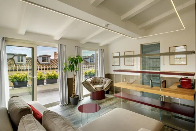 A living area with a sofa, armchair and glass shelving, with sliding doors opening onto a terrace with potted plants and rooftops beyond.