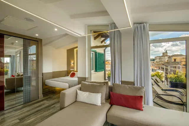 A living area with a sofa and a mirrored wardrobe, with a bed in the background, and a balcony with loungers overlooking rooftops and buildings.