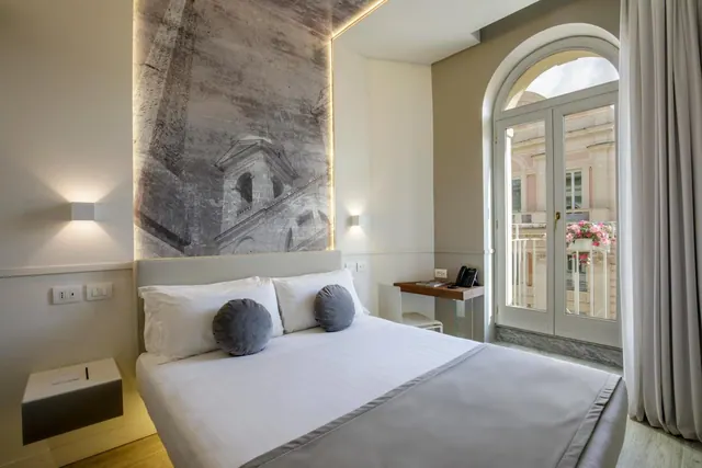 A bedroom with a double bed under a large grey mural headboard, a small wooden desk with a telephone, and arched French doors opening onto a balcony.