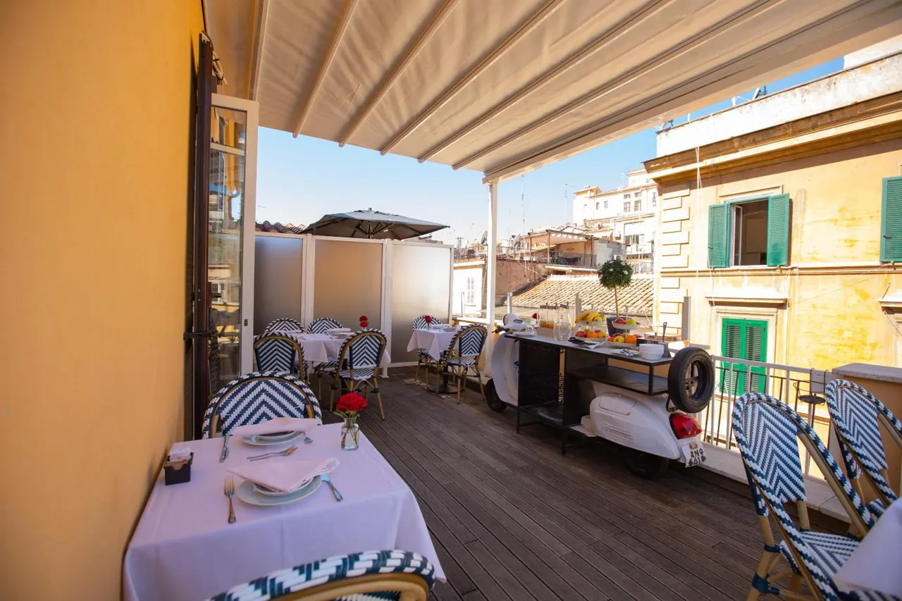 A covered rooftop terrace with laid dining tables, blue-striped chairs, a white vintage scooter holding fruit, and neighbouring buildings beyond.