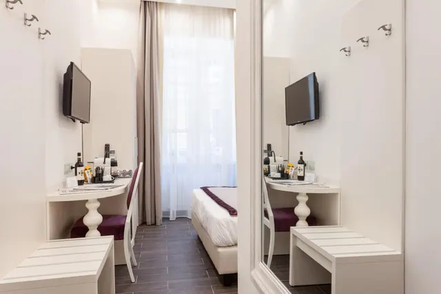A narrow hotel room with a desk and wall-mounted television on the left, a bed at the far end beside a window with sheer curtains, and a mirror reflecting the scene on the right.