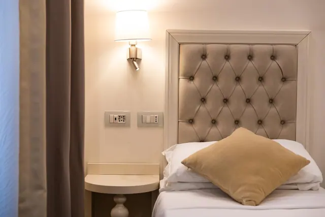 A bedroom with a single bed under a tufted upholstered headboard, a wall-mounted reading lamp, a round nightstand and taupe curtains.