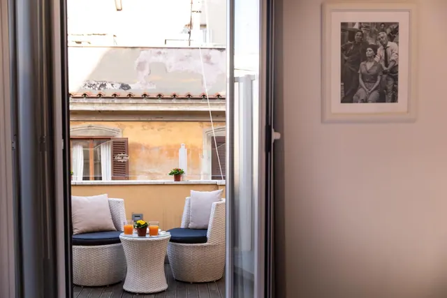 A small balcony with two wicker armchairs with navy cushions and a round wicker table holding two glasses of orange juice and a flower pot, seen through an open glass door onto the ochre wall of a neighbouring building.