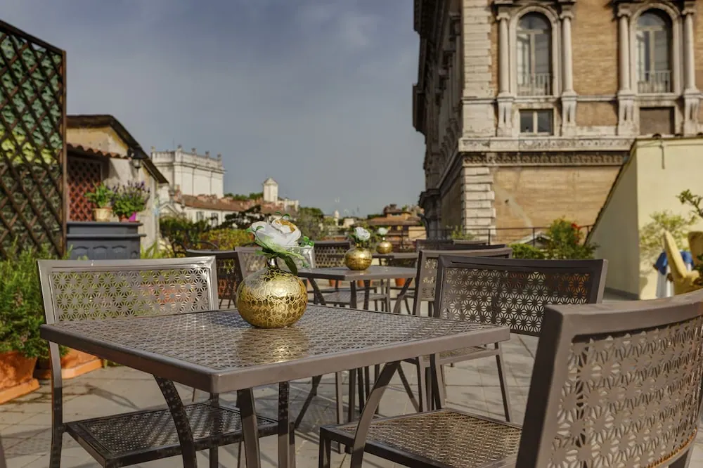 A rooftop terrace with metal tables and chairs and gold vases with white flowers, beside an ornate historic building with arched windows and rooftops visible beyond.