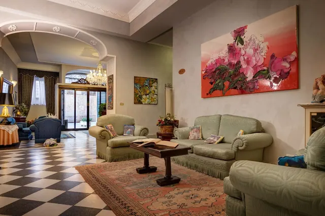 A hotel lounge with green upholstered sofas and an armchair, a checkered floor, a patterned rug, and a large pink floral painting on the wall.