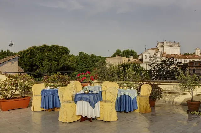 A rooftop terrace with several tables set with blue cloths and yellow chair covers, potted plants, and a view over rooftops and a white church.