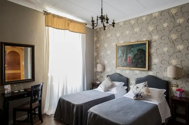 A twin bedroom with two single beds with grey covers, a wall mirror, a writing desk with a chair, a chandelier and a framed painting on floral wallpaper.
