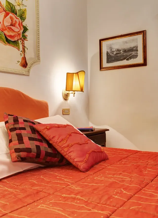 A bedroom with an orange quilted bed, patterned cushions, a lit wall lamp, a framed print and a painted rose mural on the wall.