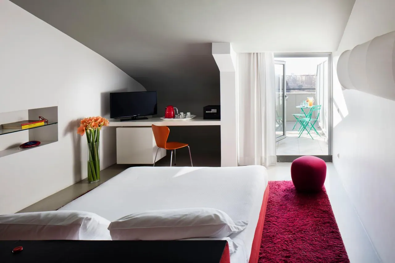 A bedroom with a double bed, a red shag rug, a desk with a television and kettle, and glass doors opening onto a balcony with teal chairs.