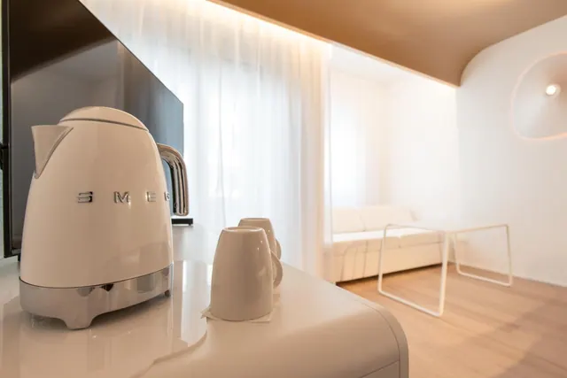 A white Smeg kettle and two cups sit on a white counter in a room with a sofa and sheer curtains.