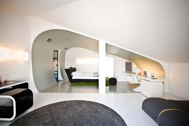 A modern hotel suite with curved white walls shows a bed in an alcove on a green rug, a white desk with a chair and a television, and a dark sofa in the foreground.