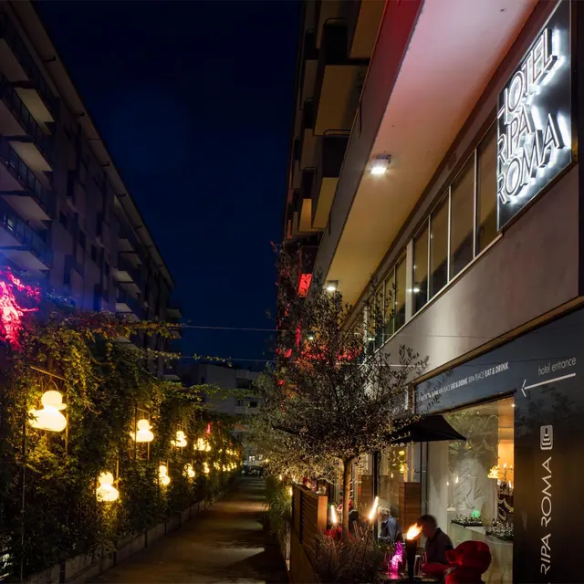 Night view of the Hotel Ripa Roma building facade with an illuminated rooftop-to-ground sign, a lit pedestrian lane, and outdoor tables with torches.
