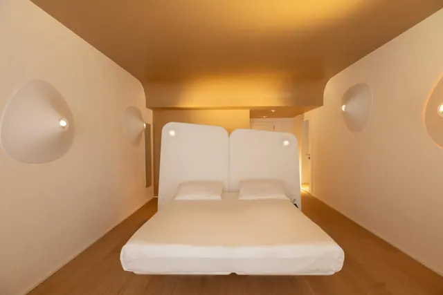 A bedroom with a double bed set against a white headboard, wall-mounted sconces, and a wooden floor under warm lighting.
