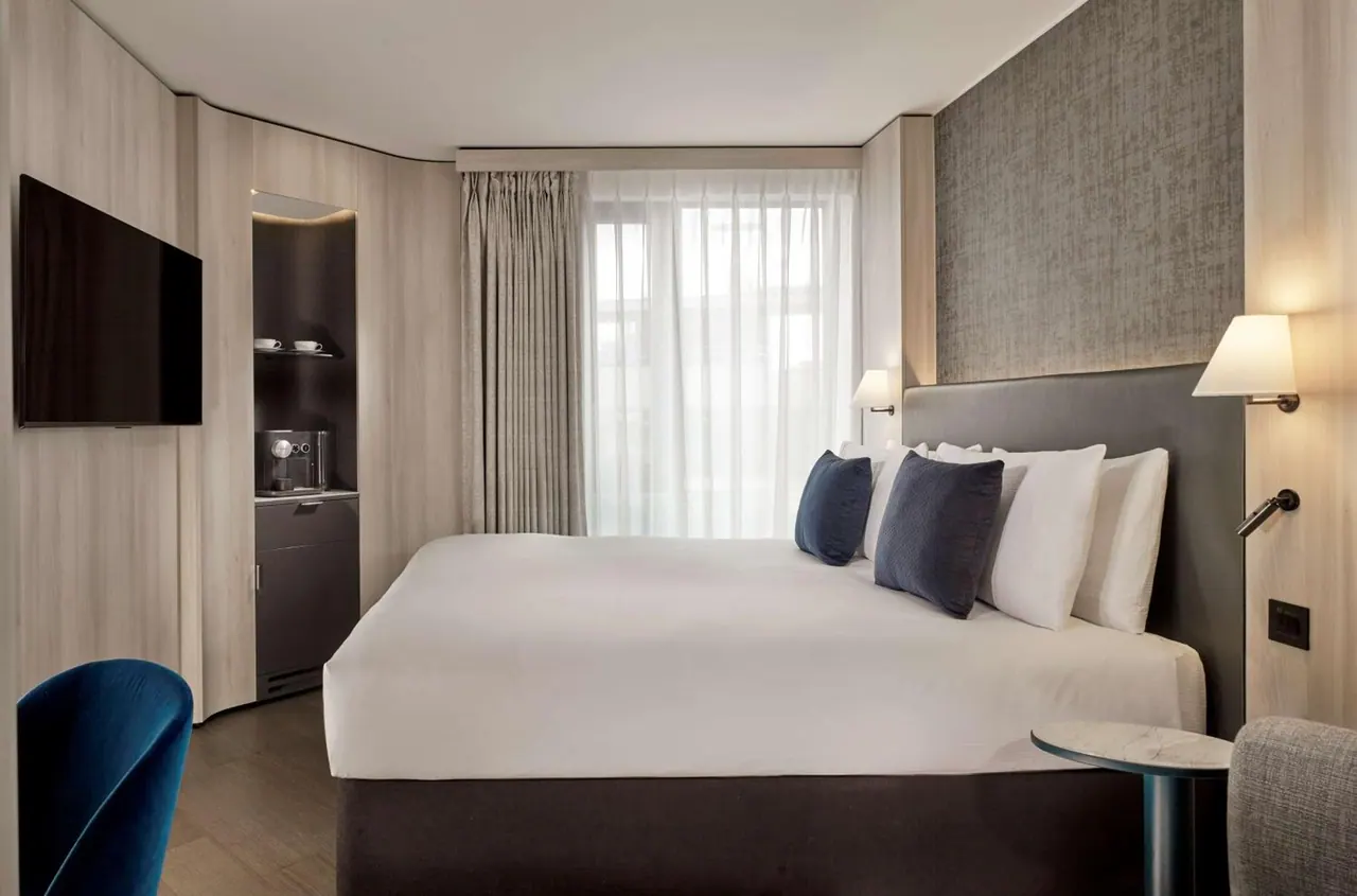 A hotel bedroom with a made double bed, a wall-mounted television, a coffee machine on a cabinet and a blue armchair in the foreground.
