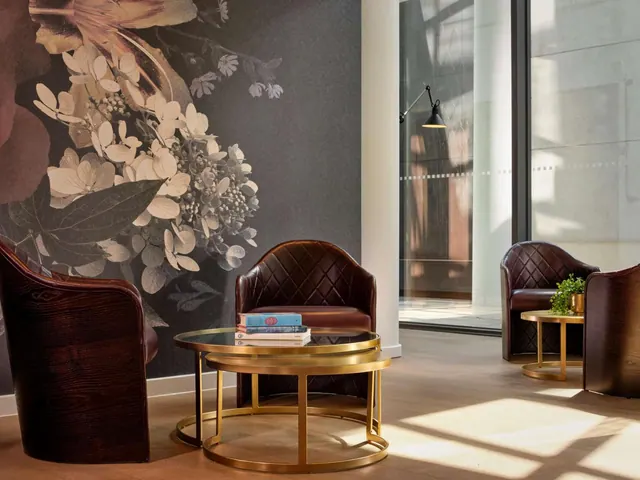 A lounge seating area with leather tub armchairs, gold nesting coffee tables holding books, a large floral wall mural and tall glass windows.