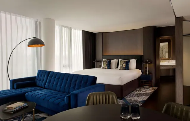 A hotel suite with a navy velvet sofa, an arc floor lamp, a king bed and a view through an open doorway into the bathroom.