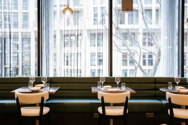 An empty restaurant dining area with green banquette seating, set tables with white upholstered chairs, and tall windows looking out onto office buildings.
