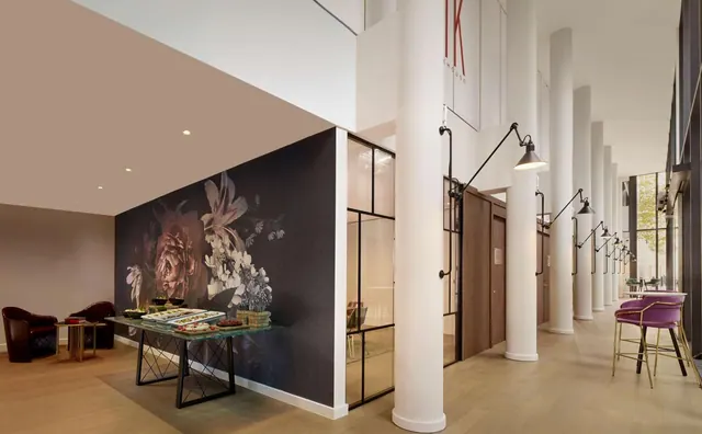 A lobby with white columns, a dark floral wall mural, a table with food platters, leather armchairs on the left and purple bar stools along the right.