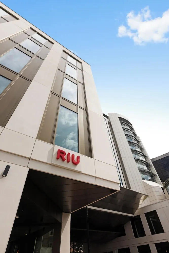 Low-angle view of a cream-coloured building facade with a red RIU sign above the entrance canopy, under a blue sky with clouds.