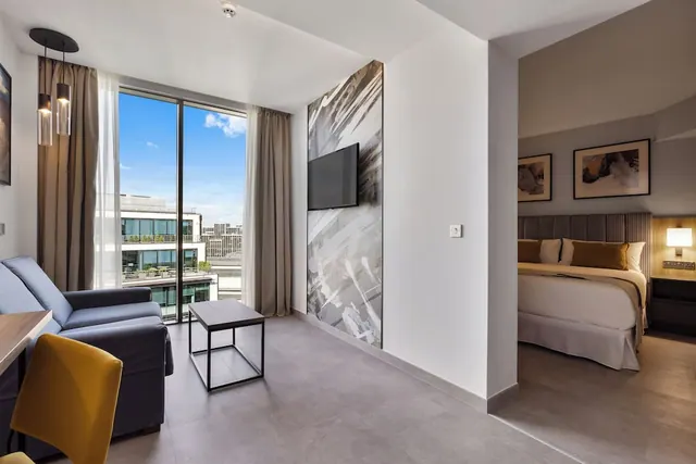 A living area with a grey sofa, yellow chair, coffee table and wall-mounted TV, with floor-to-ceiling windows overlooking buildings, and a bed visible through the doorway to the bedroom.
