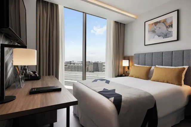 A modern hotel bedroom with a double bed, a wooden desk, a wall-mounted television and a floor-to-ceiling window looking out over distant city buildings.