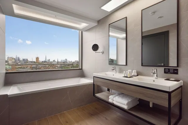 A bathroom with a long bathtub beside a large window showing a London skyline, with a double vanity and open shelving holding folded towels.
