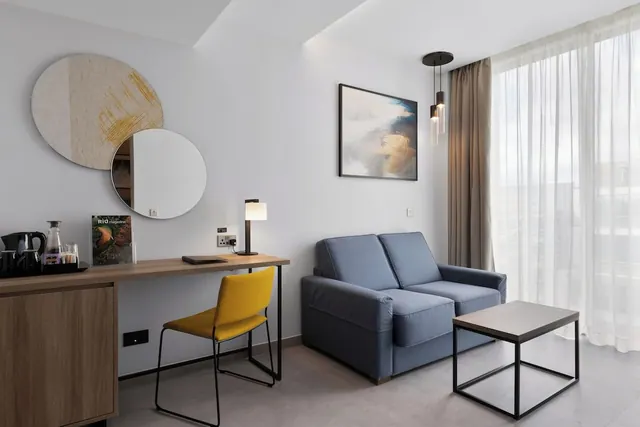 A hotel room with a wooden desk, a yellow chair, a blue two-seat sofa, a low coffee table and a window with sheer curtains.