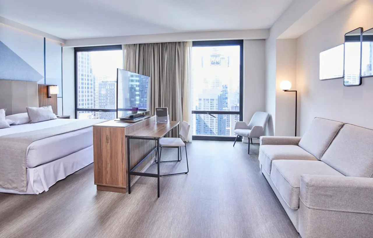 A hotel room with a king bed, a wooden desk with a flat-screen TV, a grey sofa and armchair, and large windows showing city buildings.