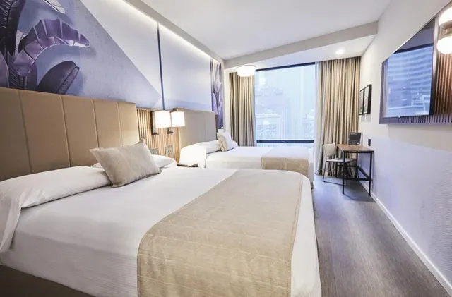 A hotel bedroom with two beds, a purple leaf mural on the wall, a wall-mounted television, a work desk, and a window looking out onto city buildings.