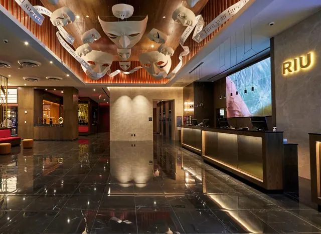 The lobby has a polished dark marble floor, a reception desk with an illuminated RIU sign on the wall, and a ceiling display of white masks.