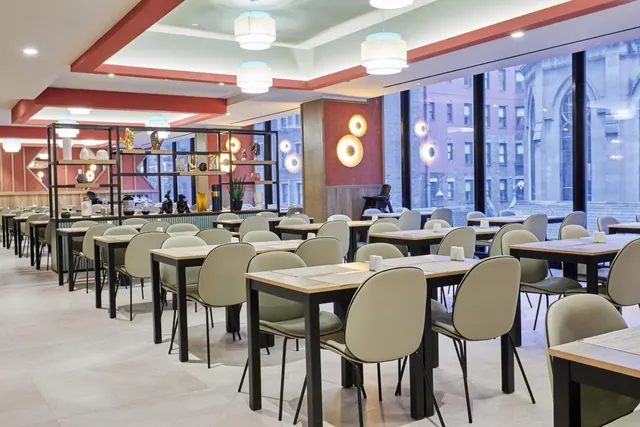 A dining room with rows of tables and pale green chairs, with floor-to-ceiling windows showing brick buildings outside.