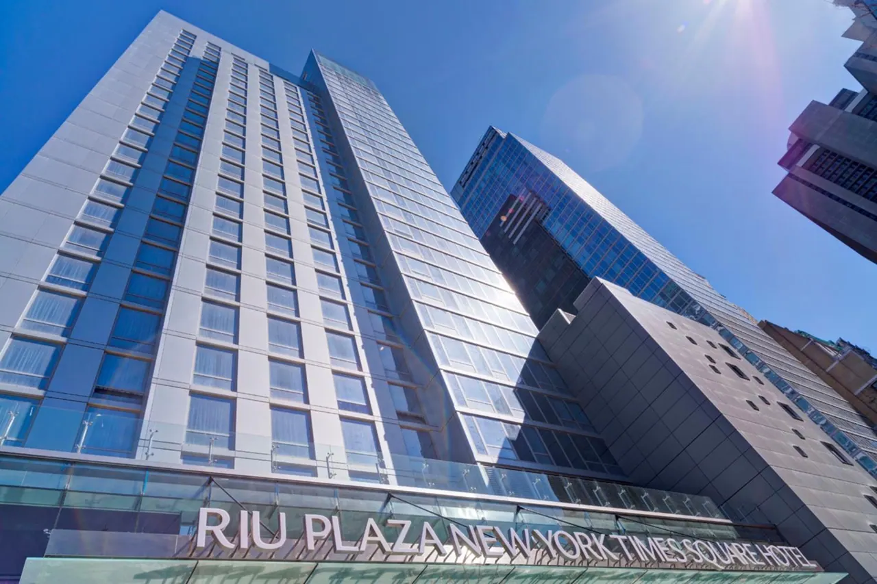 Low-angle view of a tall glass-and-white hotel tower with the sign RIU Plaza New York Times Square Hotel on its base, under a clear blue sky.