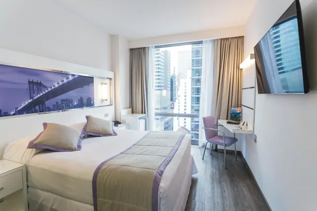 A hotel bedroom with a double bed, a bridge print on the headboard, a desk with a purple chair, a wall-mounted television, and a window looking out onto high-rise towers.