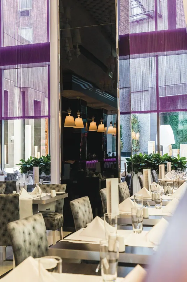 A dining room with tables set with white napkins and glassware, grey upholstered chairs, and tall windows with purple blinds.