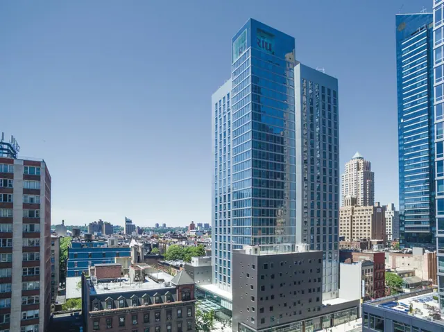 An elevated view of a tall glass hotel tower with a rooftop sign, surrounded by other city buildings under a clear blue sky.