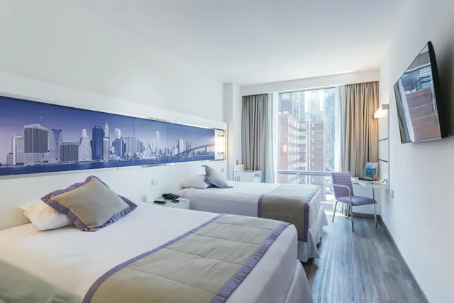A hotel room with two twin beds, a skyline mural above the headboards, a wall-mounted television and a desk by a window looking out over city buildings.