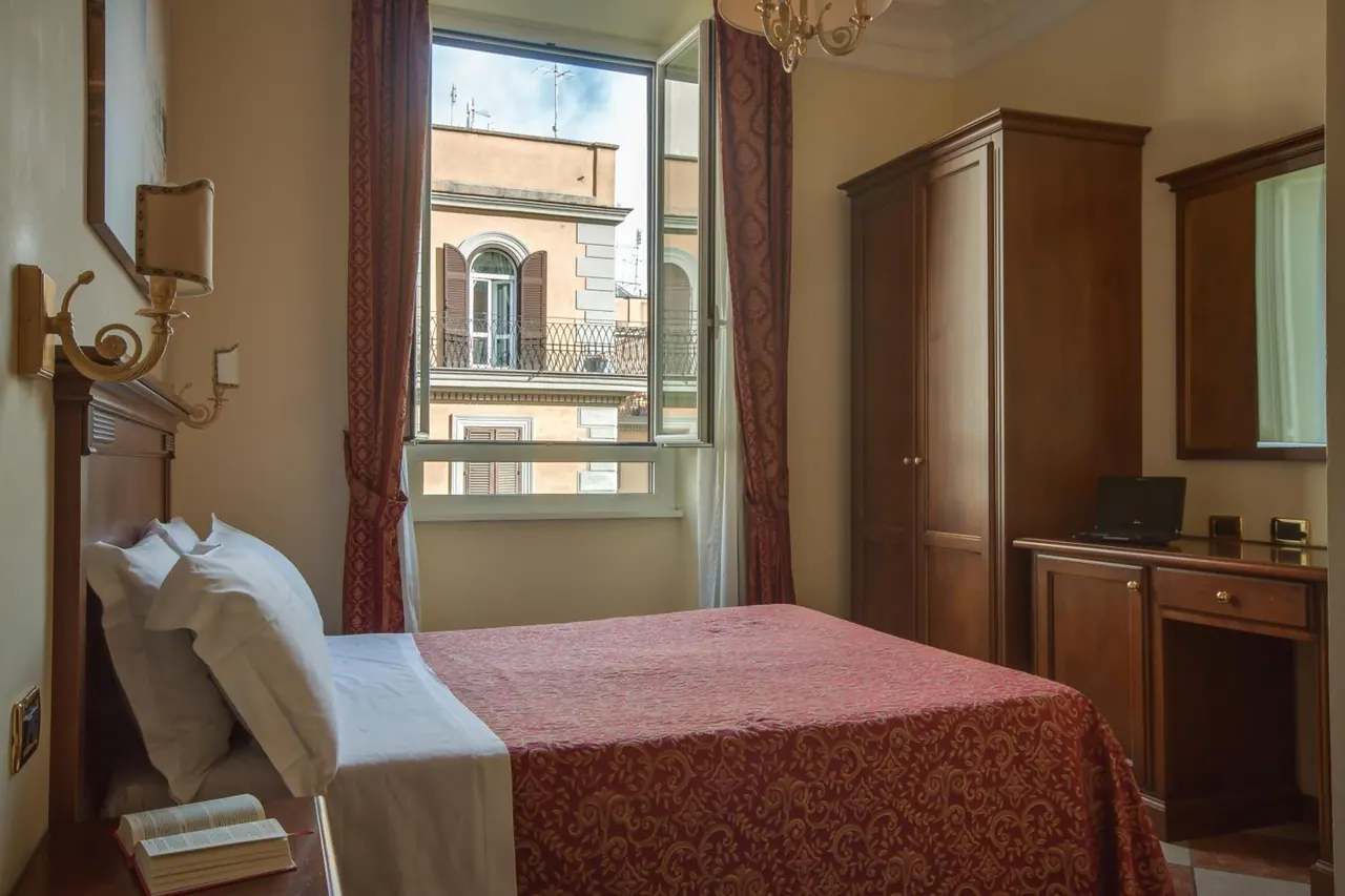 A bedroom with a made double bed, a wooden wardrobe, a desk with a laptop, and an open window looking onto a neighbouring building with a balcony.