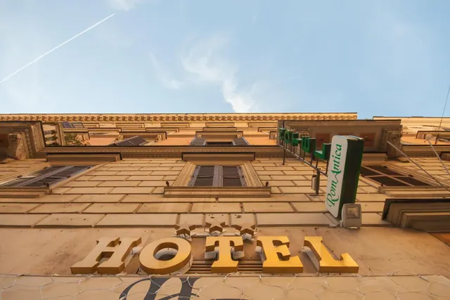 Upward view of a cream-coloured hotel facade with a gold HOTEL sign, a green Roma Antica sign and a contrail in the blue sky.