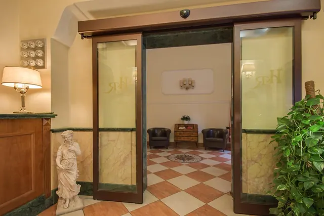 Glass sliding doors with a monogram open into a lobby lounge with two leather armchairs and a wooden chest on a checkered floor, beside a reception desk with a lamp and a statue.