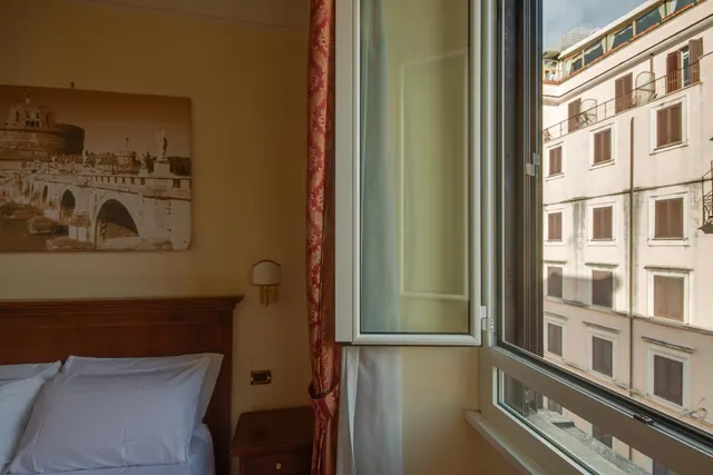 A hotel bedroom with a made bed and a framed print of Castel Sant'Angelo on the wall, with an open window looking onto a neighbouring building.