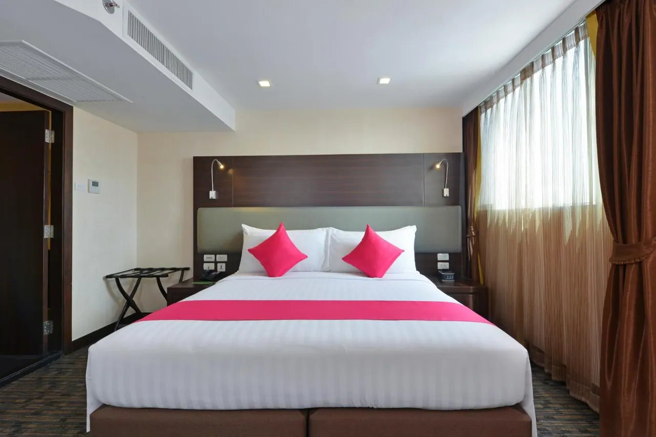 A double bed with white linens, a pink runner and pink cushions sits in a hotel bedroom beside a tall window with sheer curtains.