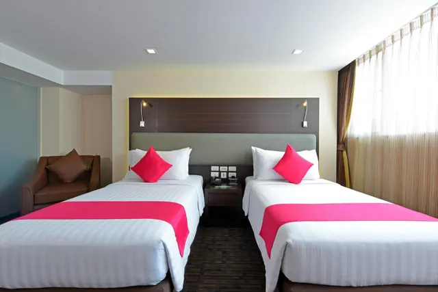A hotel bedroom with two twin beds dressed in white linen and pink runners, a brown armchair on the left and a window with sheer curtains on the right.