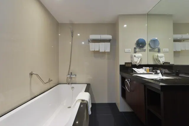 A bathroom with a white bathtub, a handheld shower over the tub, a grab rail, folded towels on a wall rack, and a vanity with a large mirror and magnifying mirrors.