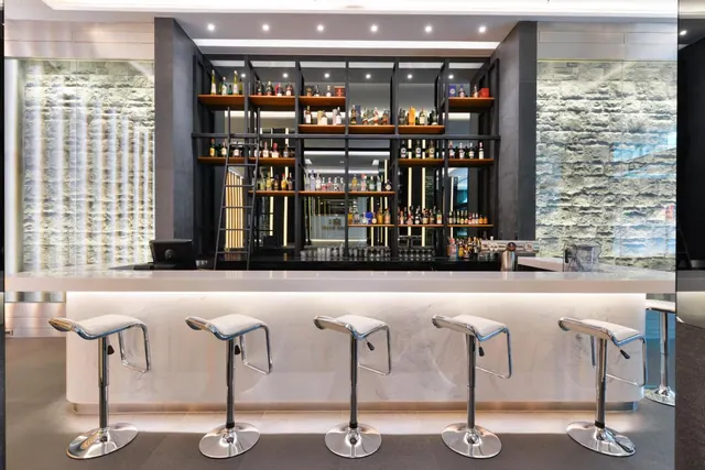 A bar counter with white swivel stools sits in front of shelves of bottles and a stone-clad wall.